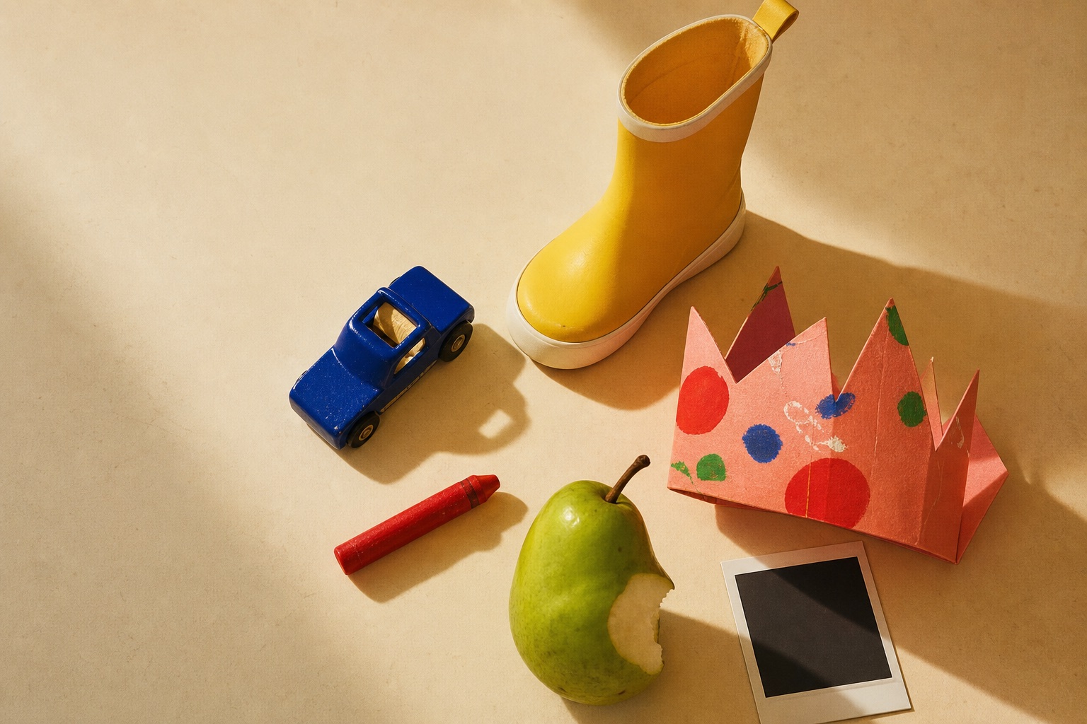 A yellow rain boot, blue toy car, paper crown, crayon, pear, and instant photo arranged on a sunlit table